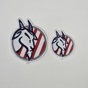 May include: Two embroidered patches featuring a goat head in profile. The goat is white with black details, set against a red and white striped background within a circular frame. The patches are in two sizes.