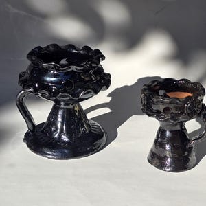 May include: Two black ceramic candle holders with a glossy finish. Each piece features a ruffled, scalloped rim, a single side handle, and a pedestal base. The larger holder has a wider bowl, while the smaller one is more compact in size.