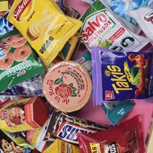 May include: An assortment of packaged snacks and food items, including chips, donuts, and candy. The image features a variety of colorful packaging, with brands like Sabritas, Takis, and Snickers visible. A round container with a rose design is also present.