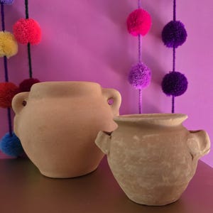 May include: Two unglazed terracotta pots with rounded bodies and small side handles. The larger pot on the left has a smooth, light tan surface, while the smaller pot on the right features a slightly textured, earthy brown finish with a wide rim.