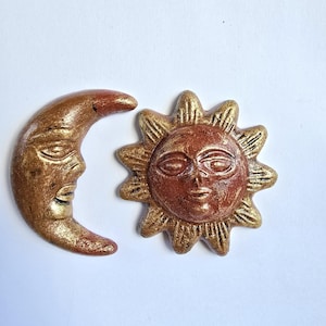 Puede incluir: Un conjunto decorativo con temática celestial, que presenta una luna creciente y un sol, ambos con caras. La luna es dorada con detalles rojos, y el sol es dorado con un centro marrón rojizo.