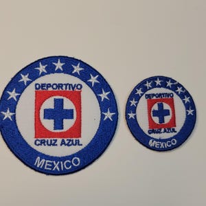 Peut inclure: Deux écussons brodés avec le logo Cruz Azul. Le plus grand est circulaire avec une bordure bleue, des étoiles blanches et les mots "DEPORTIVO CRUZ AZUL MEXICO". Le plus petit a le même design.