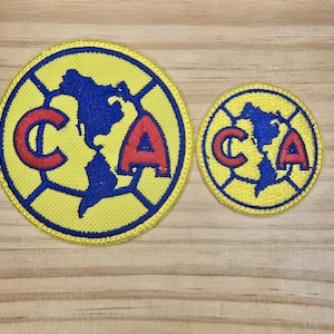 May include: Two embroidered patches featuring the letters "CA" in red with a blue and white outline. The patches are in the shape of a soccer ball with a stylized map of North and South America in the center. The larger patch is yellow with a blue border, while the smaller patch is yellow with a blue border.