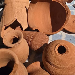 May include: A collection of terracotta pottery pieces, including several intact small vases with narrow necks and rounded bodies, alongside broken ceramic shards and fragments with a rough, matte, reddish-brown clay texture and visible cracks.