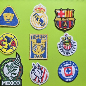May include: A collection of nine embroidered fabric patches featuring various soccer team logos, including Pumas, Real Madrid, FC Barcelona, Club America, Tigres UANL, Chivas, Mexico national team, and Cruz Azul, arranged in a grid on a green background.