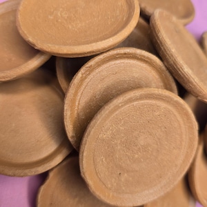 May include: A collection of small, round, terracotta-colored clay saucers. The saucers have a slightly raised edge and a smooth, unglazed surface. They are stacked and scattered, creating a textured, earthy aesthetic. The background is a soft pink.