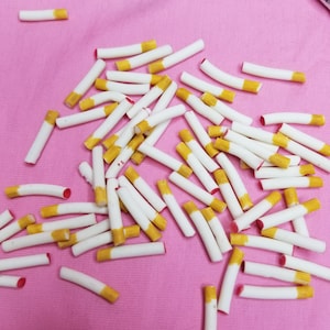 Op de afbeelding: Een verzameling witte en gele sigaretten verspreid over een roze oppervlak. De sigaretten hebben een gele filter en een rode punt. Een pakje Marlboro-sigaretten is zichtbaar op de achtergrond.