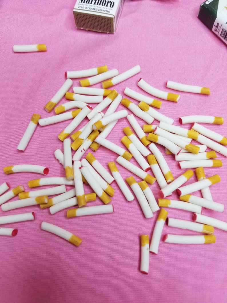 May include: A collection of white and yellow cigarettes scattered on a pink surface. The cigarettes have a yellow filter and a red tip. A pack of Marlboro cigarettes is visible in the background.