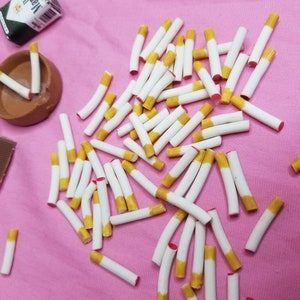 May include: Miniature cigarettes with yellow filters and white paper are scattered on a pink surface. Several packs of Marlboro cigarettes and a small ashtray are also present, creating a playful, dollhouse-like scene.