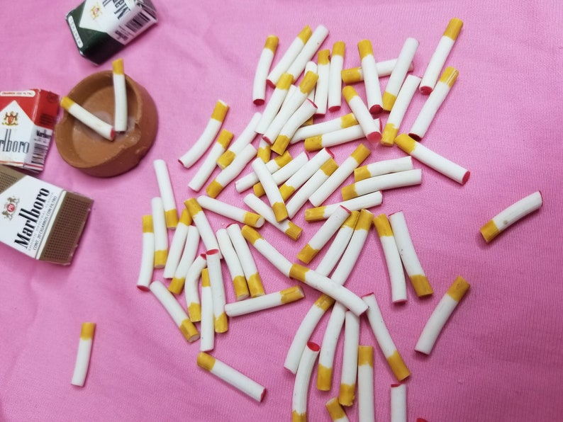 May include: Miniature cigarettes with yellow filters and white paper are scattered on a pink surface. Several packs of Marlboro cigarettes and a small ashtray are also present, creating a playful, dollhouse-like scene.