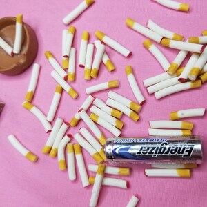 May include: A collection of white cigarettes with yellow filters scattered on a pink surface. A brown ashtray and a pack of Marlboro cigarettes are also present. An Energizer Ultimate Lithium battery is also visible.