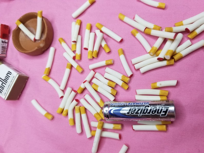 May include: A collection of white cigarettes with yellow filters scattered on a pink surface. A brown ashtray and a pack of Marlboro cigarettes are also present. An Energizer Ultimate Lithium battery is also visible.