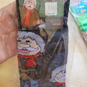 Puede incluir: Calcetines negros con un dise&ntilde;o de personaje de dibujos animados. El personaje tiene un bigote blanco, una bufanda roja y un abrigo marr&oacute;n. El embalaje dice "CALCETIN" y "DR. SIMI DR. CHAPATIN". Los calcetines son unisex.