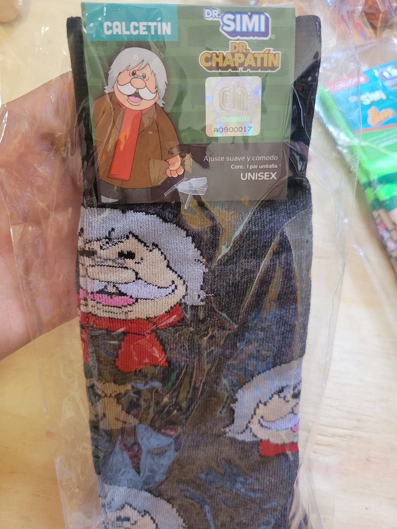 Puede incluir: Calcetines negros con un dise&ntilde;o de personaje de dibujos animados. El personaje tiene un bigote blanco, una bufanda roja y un abrigo marr&oacute;n. El embalaje dice "CALCETIN" y "DR. SIMI DR. CHAPATIN". Los calcetines son unisex.