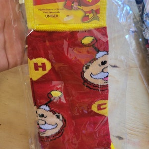 Puede incluir: Calcetines rojos con un dise&ntilde;o de personaje de dibujos animados, detalles amarillos y la letra "H". Los calcetines est&aacute;n en una bolsa de pl&aacute;stico transparente. El embalaje incluye la palabra "UNISEX".
