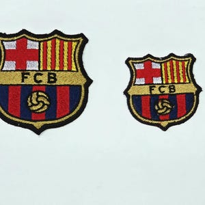 May include: Two embroidered patches featuring the FC Barcelona crest. The crests are shield-shaped with a gold border. The top section has a red cross and vertical yellow and red stripes. The bottom section is blue and red with a soccer ball.