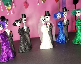 May include: A collection of five pairs of skeleton figurines, each featuring a figure in a glittery dress and a figure in a black tuxedo with a top hat. The dresses are purple, black, white, blue, and green, with matching hats or veils.
