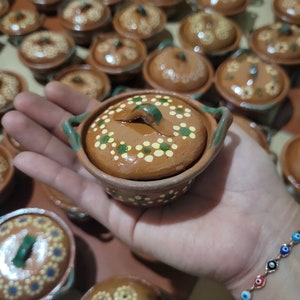Puede incluir: Una colección de pequeñas ollas de barro marrón con tapas. Las ollas están decoradas con puntos blancos y verdes. Una olla está siendo sostenida en una mano con una pulsera colorida.