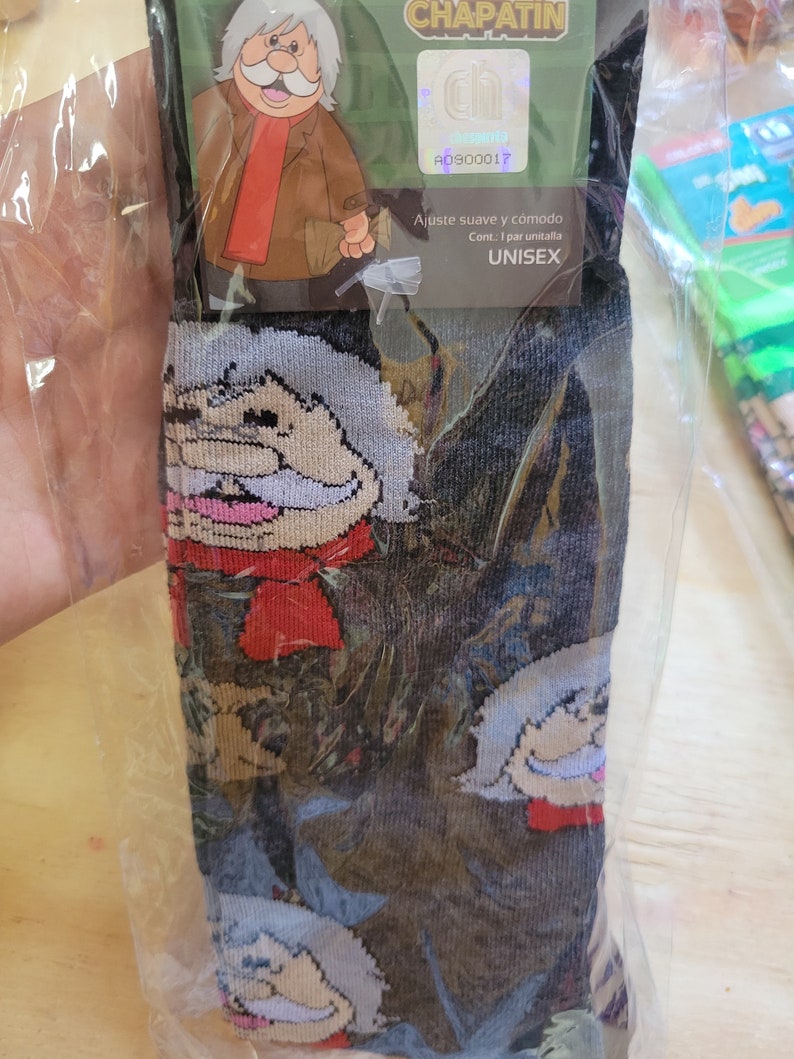 Puede incluir: Calcetines unisex con un personaje de dibujos animados. El dise&ntilde;o repetido presenta a un hombre con pelo blanco, bufanda roja y chaqueta marr&oacute;n. Los calcetines son negros con la cara del personaje repetida. El embalaje incluye el texto "CHAPATIN".