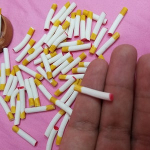May include: A collection of miniature cigarettes with white tubes, yellow filters, and red tips, scattered on a pink surface. A small, brown, terracotta bowl is also present. The image suggests a craft or dollhouse accessory.