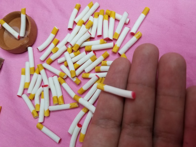 May include: A collection of miniature cigarettes with white tubes, yellow filters, and red tips, scattered on a pink surface. A small, brown, terracotta bowl is also present. The image suggests a craft or dollhouse accessory.