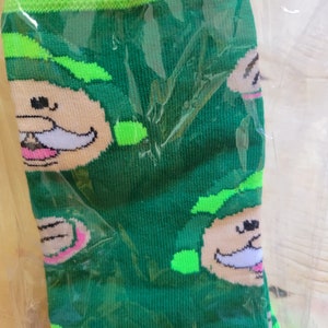 Puede incluir: Calcetines verdes con un patr&oacute;n repetido de una cara de duende de dibujos animados con bigote blanco y boca rosa. La parte superior y la puntera de los calcetines son de color verde brillante. Los calcetines est&aacute;n en un embalaje de pl&aacute;stico transparente.