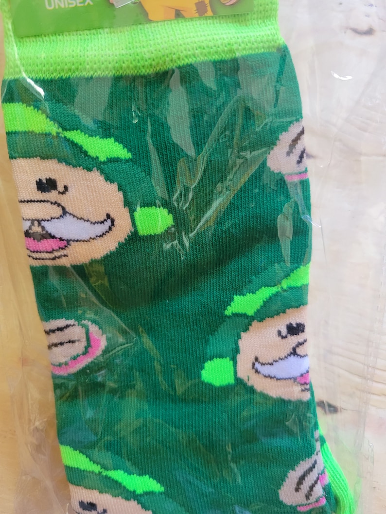 Puede incluir: Calcetines verdes con un patr&oacute;n repetido de una cara de duende de dibujos animados con bigote blanco y boca rosa. La parte superior y la puntera de los calcetines son de color verde brillante. Los calcetines est&aacute;n en un embalaje de pl&aacute;stico transparente.