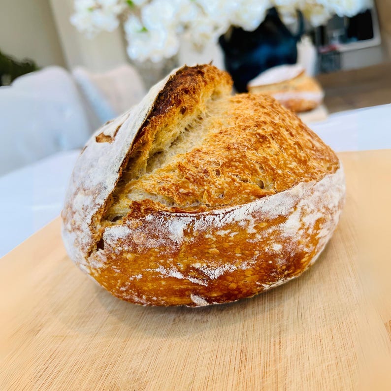 May include: A freshly baked loaf of sourdough bread, resting on a wooden cutting board. The crust is golden brown with a crackled surface, dusted with white flour. The bread's interior is visible where it has been cut, showing a light, airy texture.