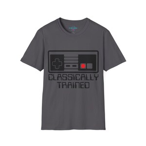 Könnte beinhalten: Dunkelgraues T-Shirt mit einem schwarzen Controller-Design und einem roten Knopf. Darunter steht in fetter, eckiger Schrift der Text "CLASSICALLY TRAINED". Das T-Shirt hat einen Rundhalsausschnitt und kurze Ärmel.