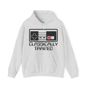 May include: A light gray hoodie featuring a black and white graphic of a classic video game controller. The text "CLASSICALLY TRAINED" is printed below the controller in black, with a kangaroo pocket and drawstring hood.