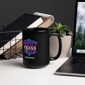 May include: Black ceramic mug with a white design that says "Protect Trans Youth" in pink and blue lettering. The design also includes a stylized compass and the word "Euphorium" in white lettering.