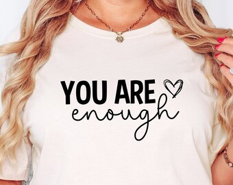 May include: Cream-colored t-shirt with the text "YOU ARE enough" in black script and a heart symbol. The shirt is a crew neck and appears to be made of a soft material. The image shows the shirt being worn.