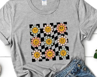 May include: A gray t-shirt featuring a retro-style design of smiling flowers on a black and white checkered background. The flowers have yellow centers and pastel-colored petals. The shirt is paired with denim shorts.