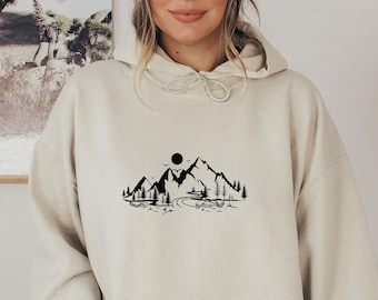 May include: A cream-colored hoodie with a black mountain range graphic and a sun above the mountains. The hoodie has a drawstring hood and a kangaroo pocket.