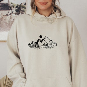 K&ouml;nnte beinhalten: Ein cremefarbener Hoodie mit einem schwarzen Bergpanorama-Motiv und einer Sonne &uuml;ber den Bergen. Der Hoodie hat eine Kapuze mit Kordelzug und eine K&auml;ngurutasche.