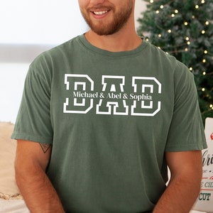 May include: A green t-shirt with the word "DAD" in white block letters. The names "Michael & Abel & Sophia" are written in smaller white letters below the word "DAD".