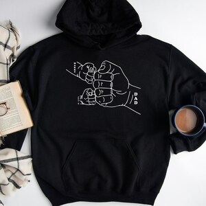 May include: Black hooded sweatshirt with a white outline of a father's hand holding the hands of three children. The text "DAD" is written on the father's wrist.