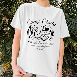 Op de afbeelding: Wit T-shirt met een zwart grafisch ontwerp. Het ontwerp toont de tekst "Camp Olivia" boven een berg- en rivierlandschap met bomen en een zon. Daaronder staat "Olivia's Bachelorette Lake Tahoe, California est. 2025".