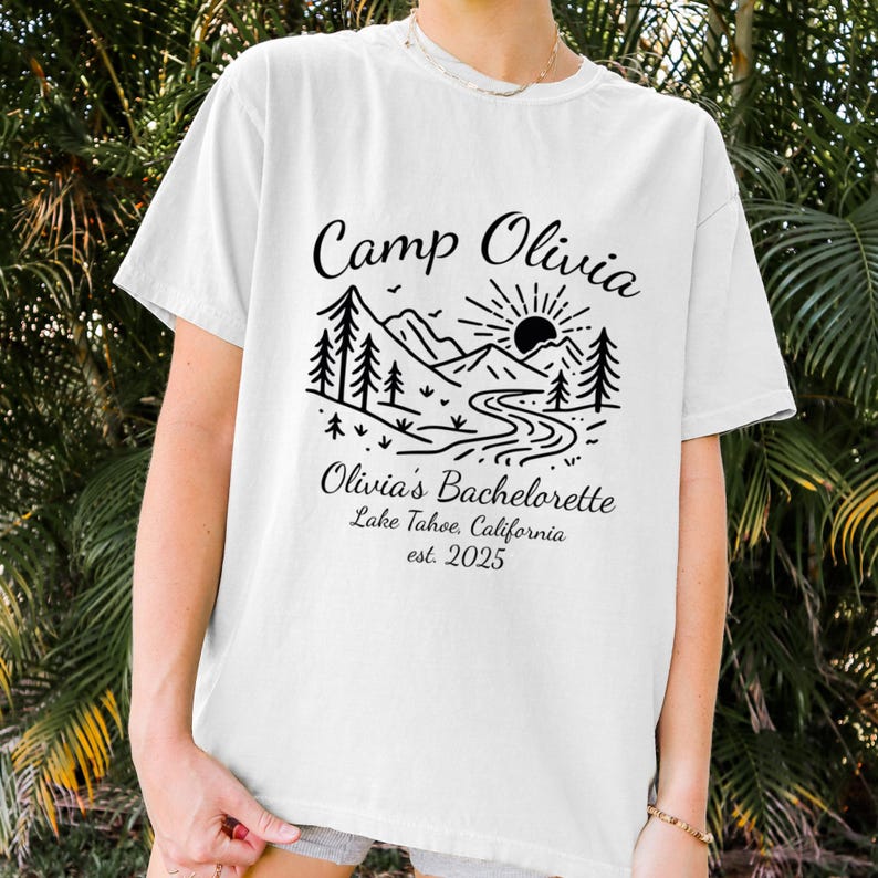 Op de afbeelding: Wit T-shirt met een zwart grafisch ontwerp. Het ontwerp toont de tekst "Camp Olivia" boven een berg- en rivierlandschap met bomen en een zon. Daaronder staat "Olivia's Bachelorette Lake Tahoe, California est. 2025".
