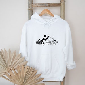 K&ouml;nnte beinhalten: Wei&szlig;er Kapuzenpullover mit einem schwarzen Aufdruck einer Bergkette mit Sonne und B&auml;umen.