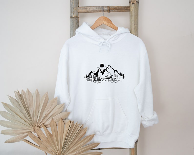K&ouml;nnte beinhalten: Wei&szlig;er Kapuzenpullover mit einem schwarzen Aufdruck einer Bergkette mit Sonne und B&auml;umen.