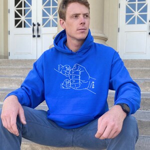 May include: A royal blue hooded sweatshirt with a white graphic of a father and child's hands clasped together in a fist bump. The text "DAD" is written on the father's arm.