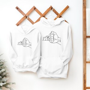 May include: Two white hooded sweatshirts with a black line drawing of two hands clasped together in a fist bump. The smaller hand is on the left and the larger hand is on the right.