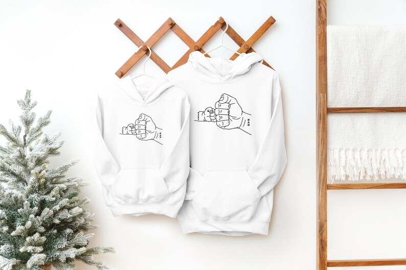 May include: Two white hooded sweatshirts with a black line drawing of two hands clasped together in a fist bump. The smaller hand is on the left and the larger hand is on the right.