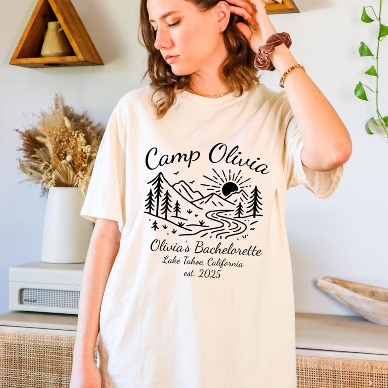 Op de afbeelding: Cr&egrave;mekleurig T-shirt met een zwart grafisch ontwerp. Het ontwerp toont de tekst "Camp Olivia" boven een berg- en rivierlandschap met een zon. Daaronder staat "Olivia's Bachelorette" en "Lake Tahoe, California est. 2025".