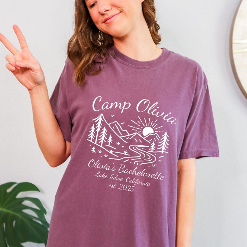 Op de afbeelding: Een mauvekleurig T-shirt met een witte grafische afbeelding. De afbeelding toont de tekst "Camp Olivia" boven een berg- en rivierlandschap met bomen en een zon. Daaronder staat "Olivia's Bachelorette Lake Tahoe, California est. 2025."