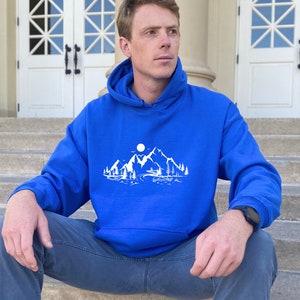 K&ouml;nnte beinhalten: Ein k&ouml;nigsblauer Kapuzenpullover mit einem wei&szlig;en Grafikdruck, der eine Bergszene zeigt. Die Grafik zeigt eine Bergkette, einen Vollmond, B&auml;ume und einen Fluss.