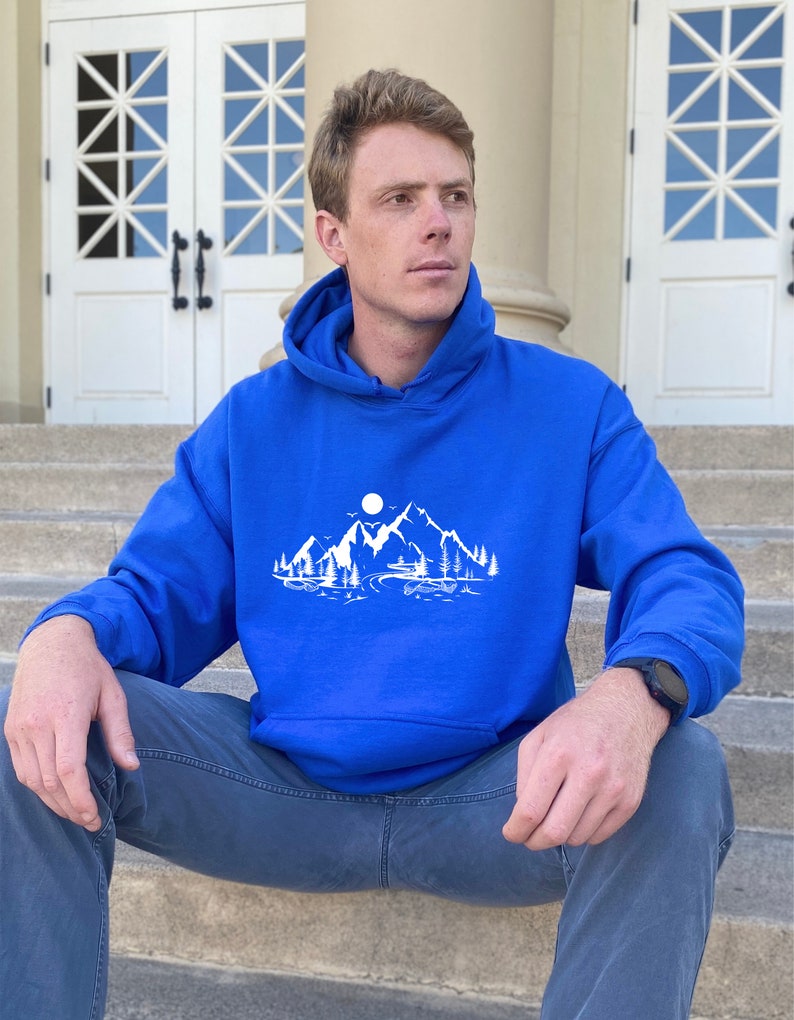 K&ouml;nnte beinhalten: Ein k&ouml;nigsblauer Kapuzenpullover mit einem wei&szlig;en Grafikdruck, der eine Bergszene zeigt. Die Grafik zeigt eine Bergkette, einen Vollmond, B&auml;ume und einen Fluss.