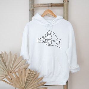 May include: White hooded sweatshirt with a black line drawing of two fists bumping, one labeled "Dad" and the other "Little."