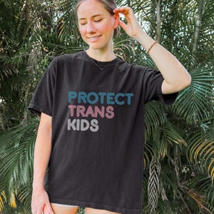 May include: Black t-shirt with the text "PROTECT TRANS KIDS" in blue, pink, and white. The shirt is a dark color and has a casual, relaxed fit. The text is stacked vertically on the front of the shirt.